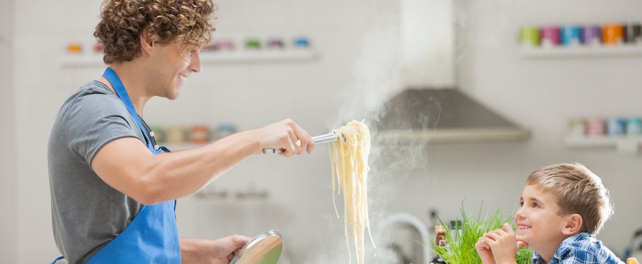 A father wearing an apron scoops spaghetti with tongs, while his son sits at the counter smiling at him.