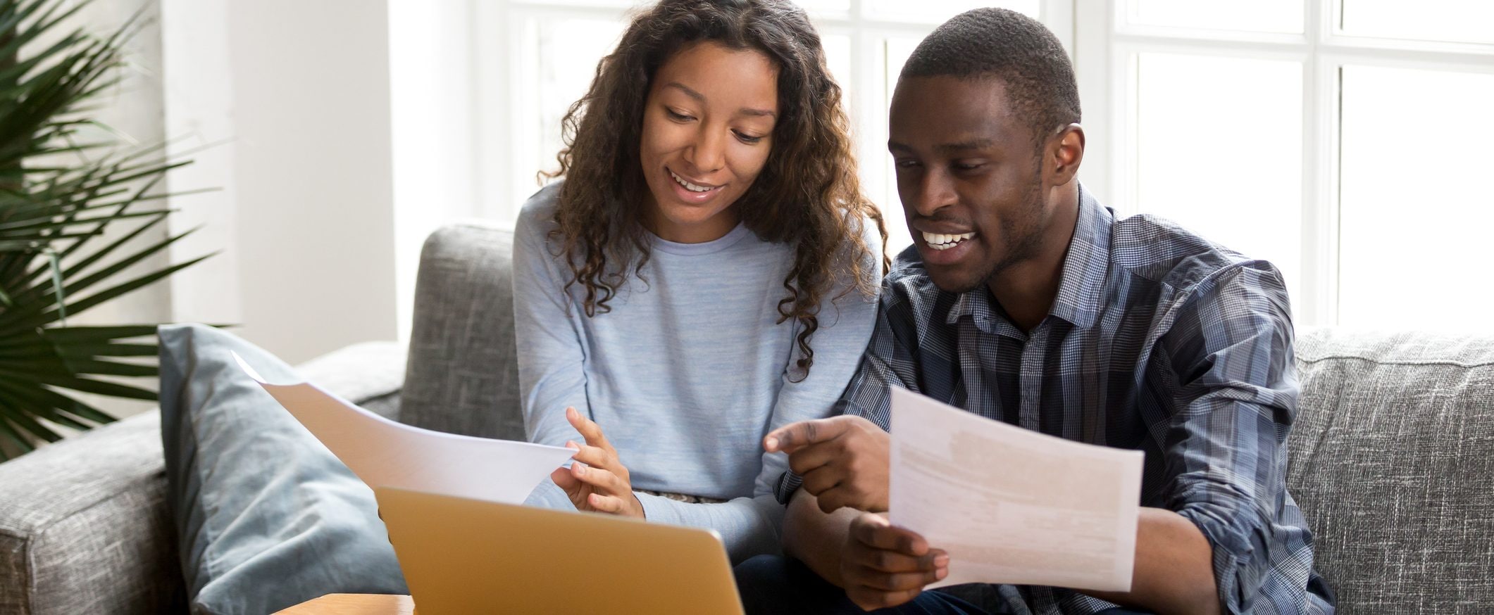 A man and a woman sit on a couch together looking over papers and at the screen of a laptop.