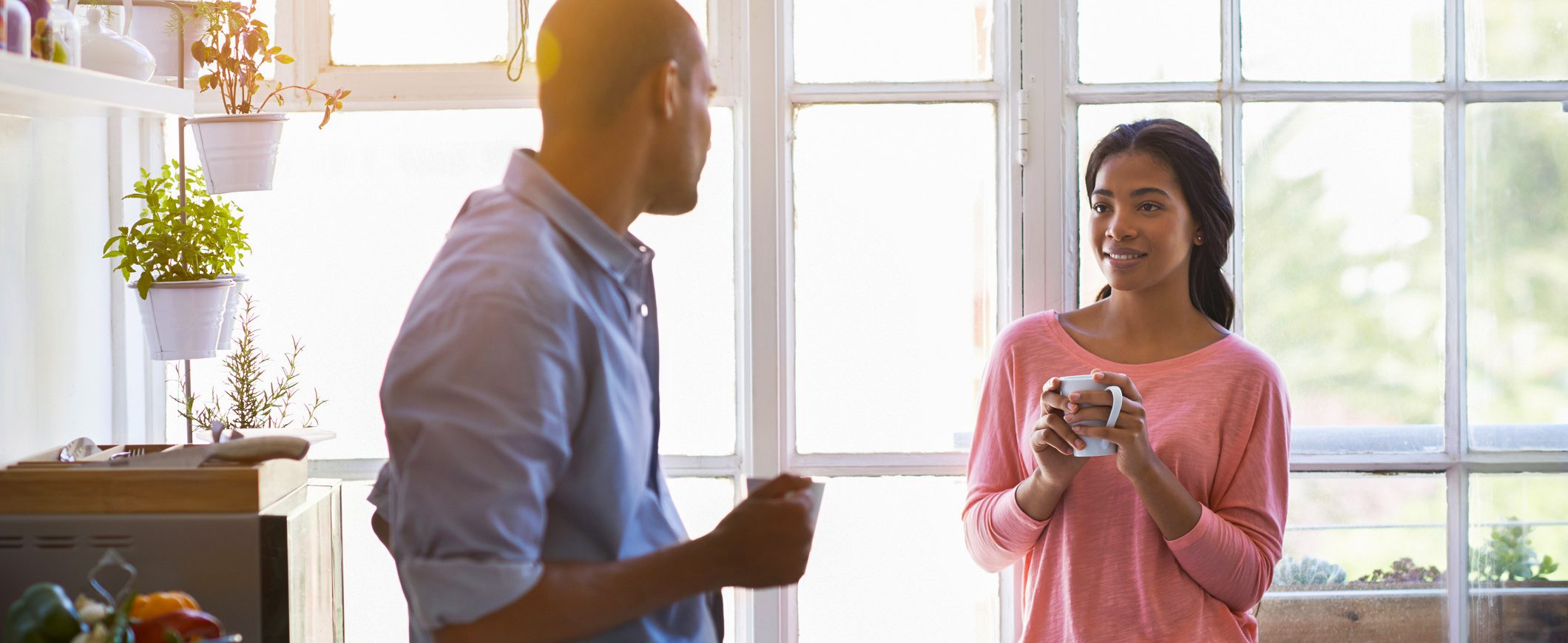 A couple holding coffee mugs looks at each other in front of the windows inside a home.