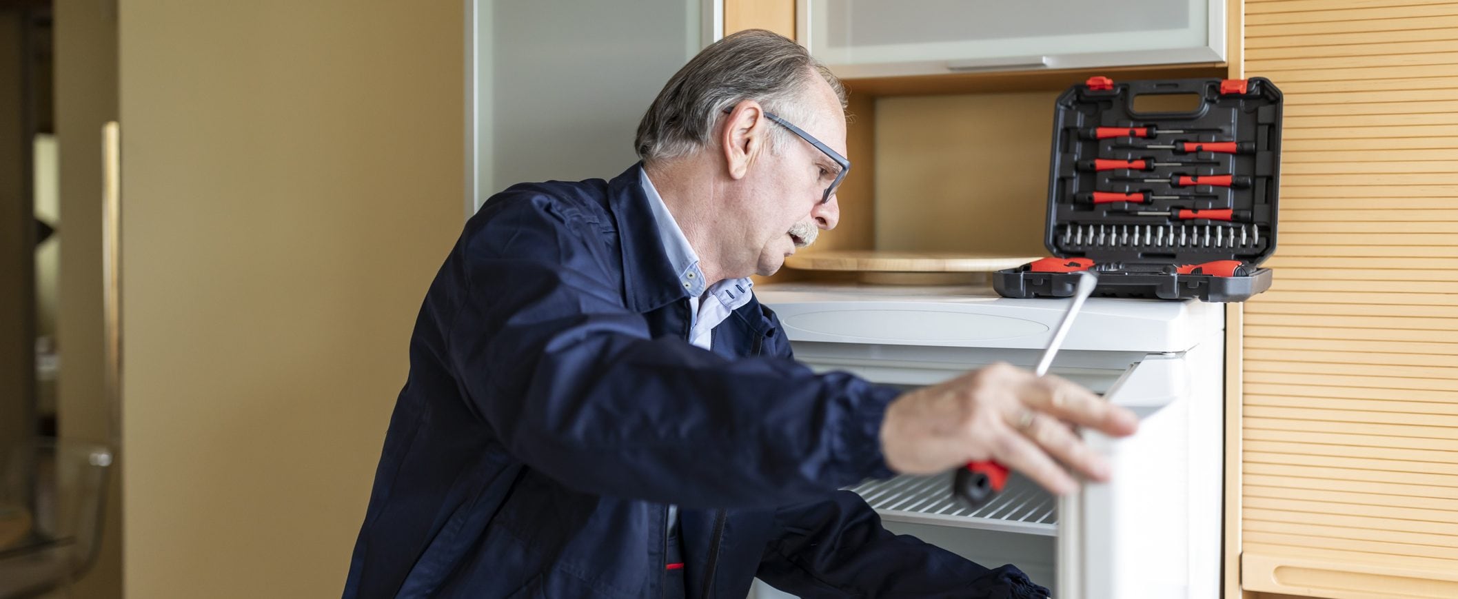 A repairman adjusts something inside a refrigerator. A toolbox sits on top of the refrigerator.