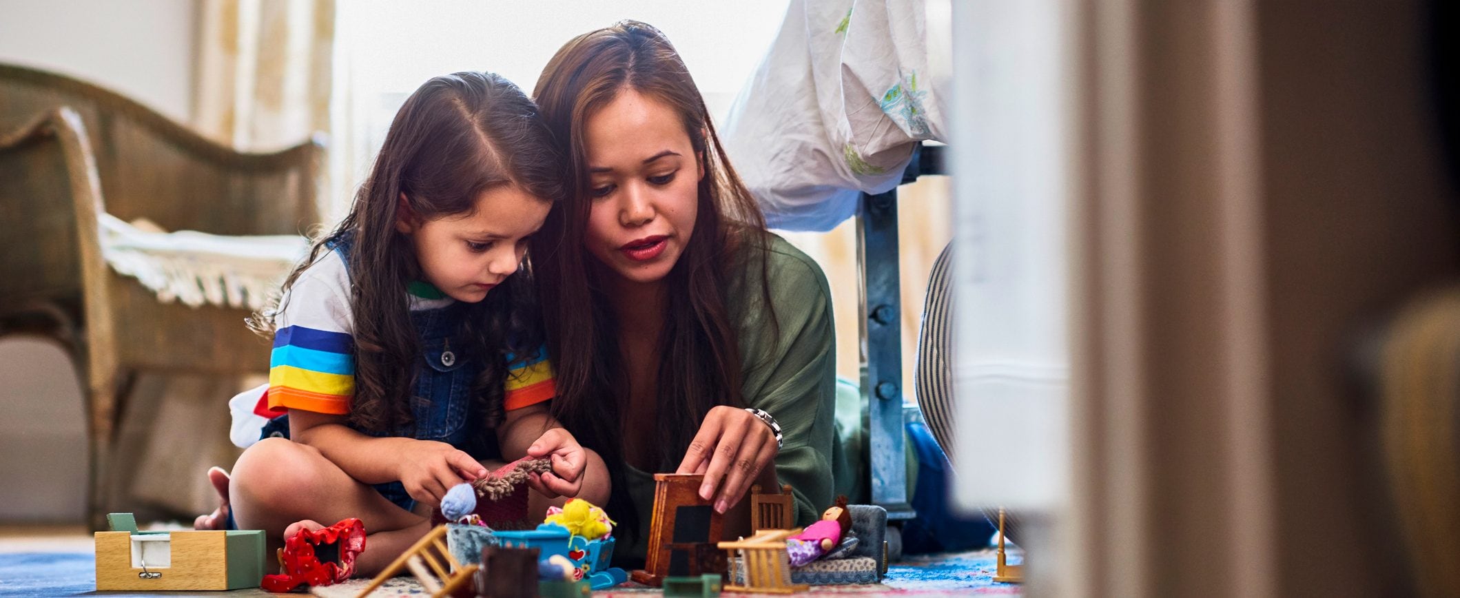 A mom and young daughter play on the floor with doll house furniture and figures.