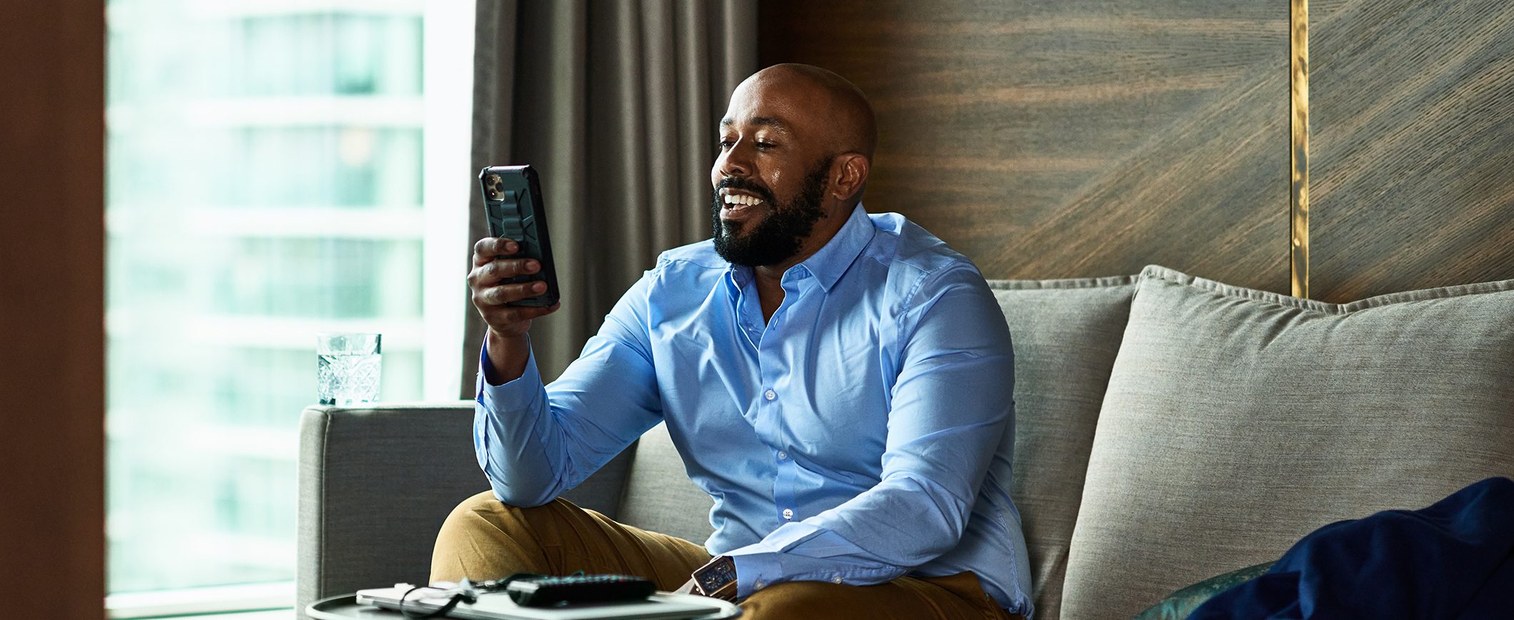 A man sits on a couch smiling at his mobile device.
