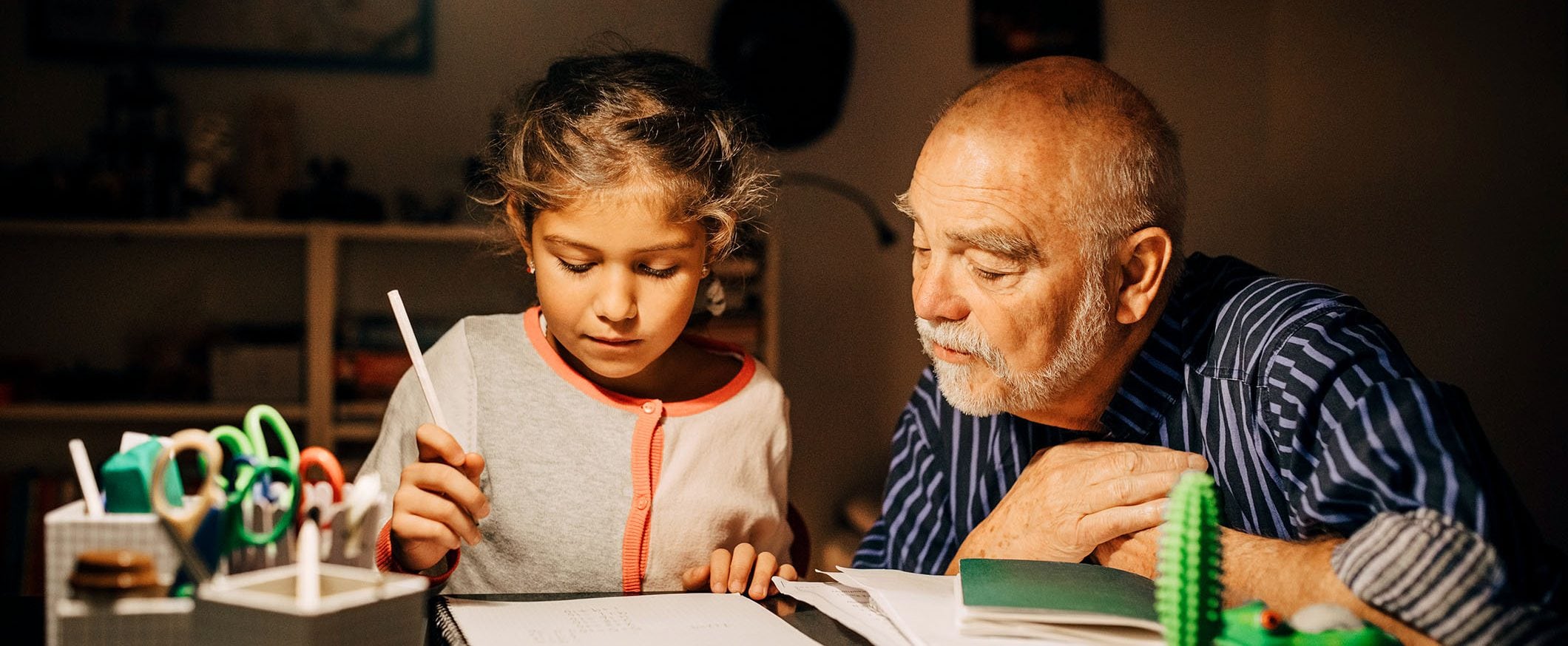 A man and young girl are seated at a table, she is holding a pen and they’re looking at a drawing.