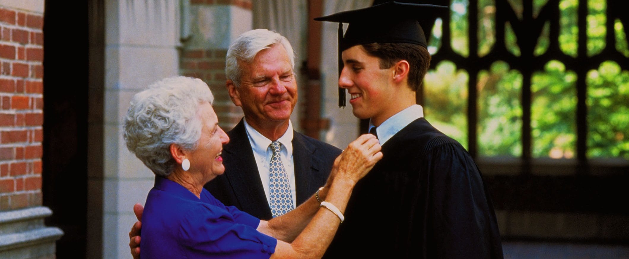 An older couple smile while standing with a young man dressed in a cap and gown, with a school in the background.