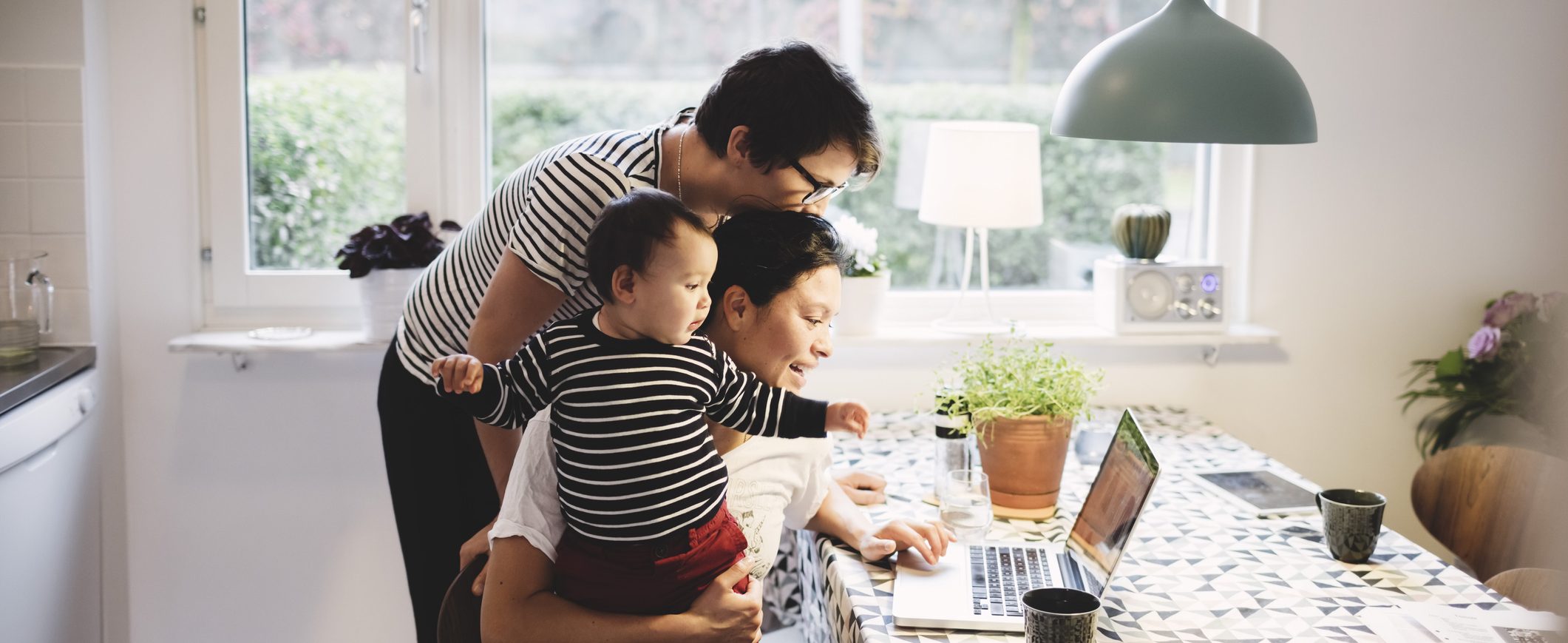 Two women look at a laptop at a kitchen table. One of them holds a baby.