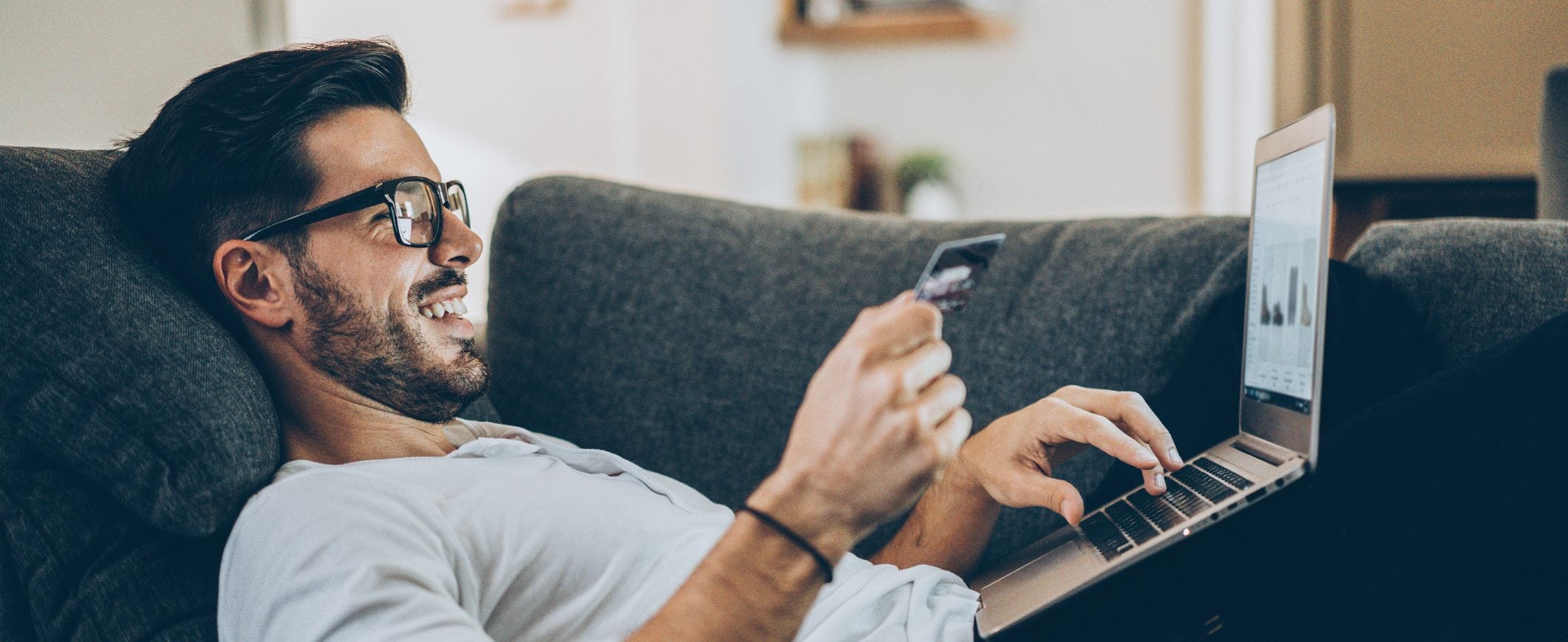 A man reclines on a couch with a laptop. He holds a credit card in one hand