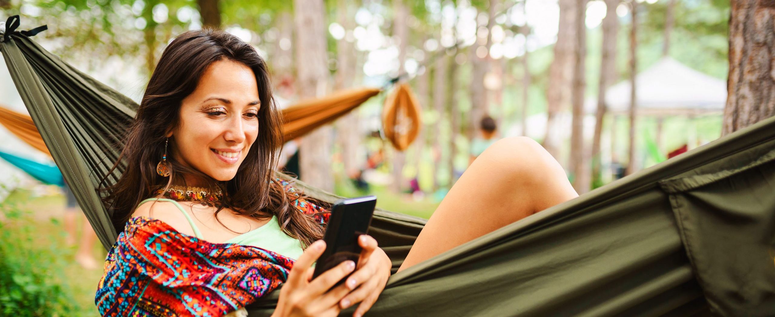 A woman smiles while reclining in a hammock and looking at her mobile phone, with trees in the background.