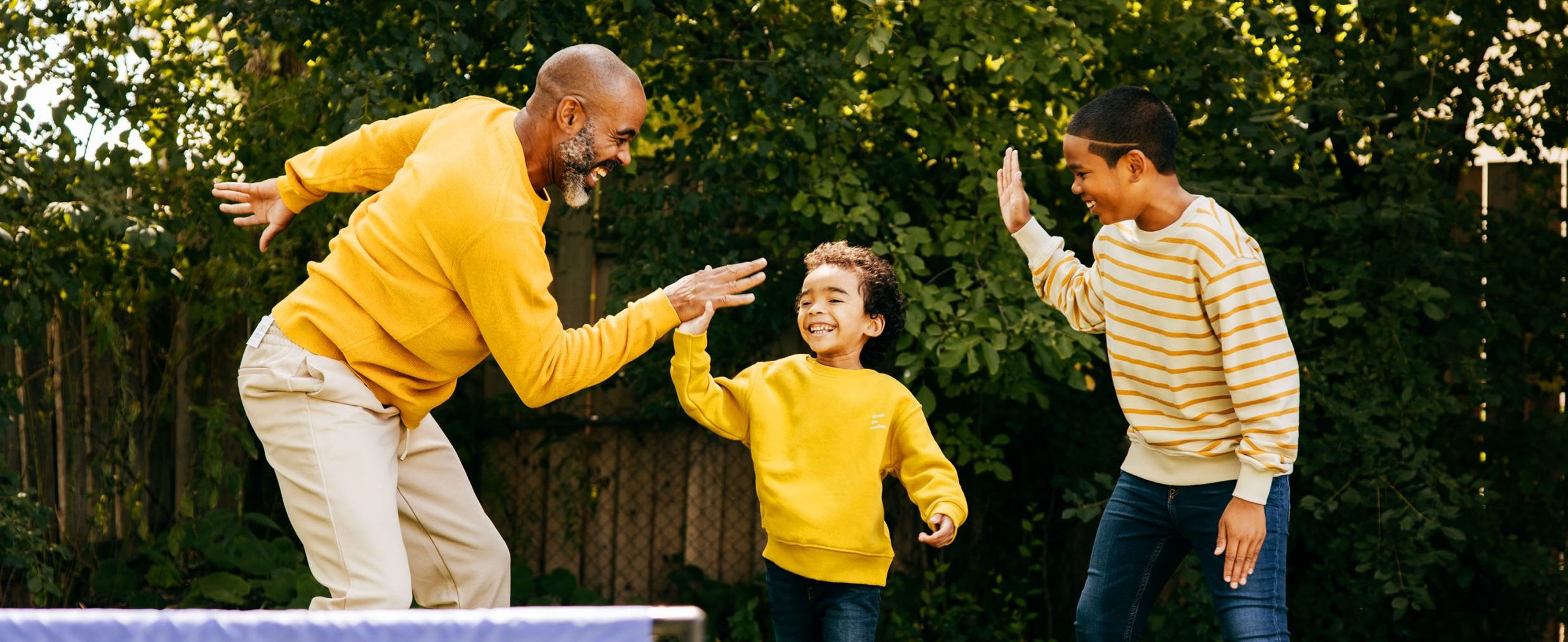 A man smiles while high-fiving two boys in a backyard, with trees in the background.