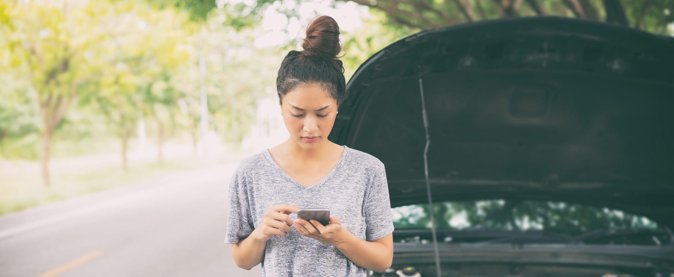 A woman stands on the side of a street in front of her car that has its hood up, looking at her smartphone.