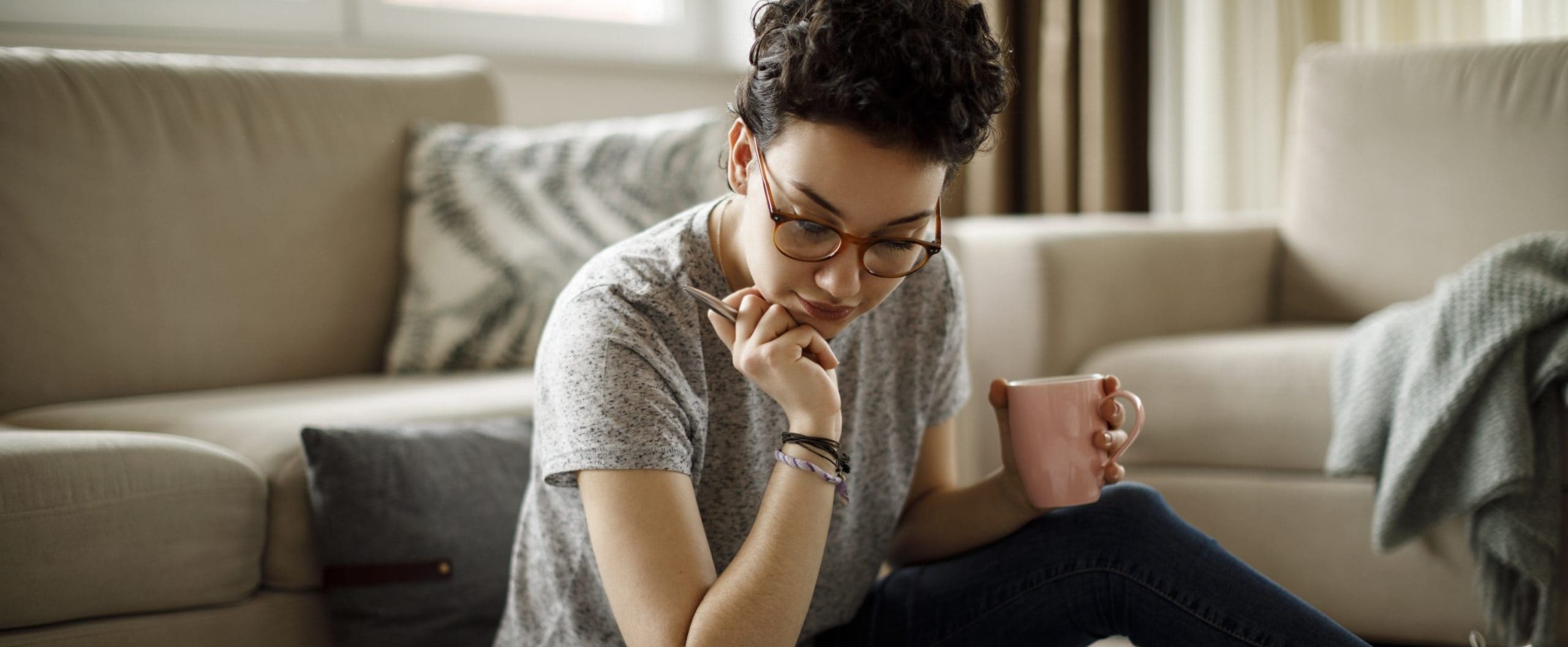 A person, holding a pen and a coffee cup, sits on the ground next to a couch looking at a laptop.