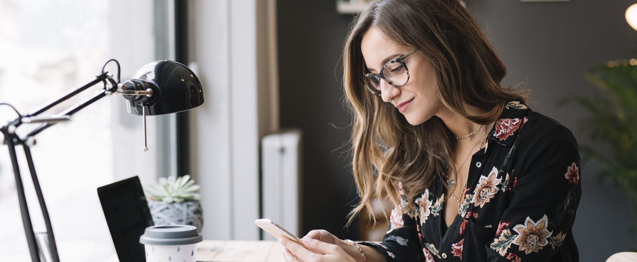 A woman, smiling, sits at her desk and checks her online savings account on her phone.