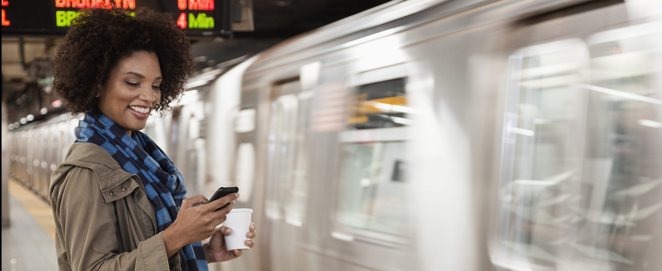 A woman waiting for her subway train and holding a coffee checks her online savings account on her phone.