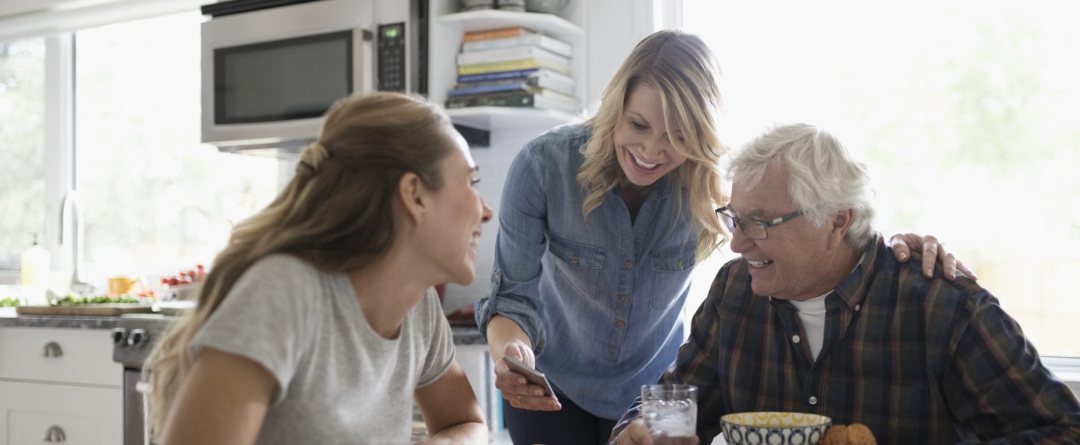 A man and a woman sitting at a kitchen table smiling, while a woman standing shows them something on a smartphone.