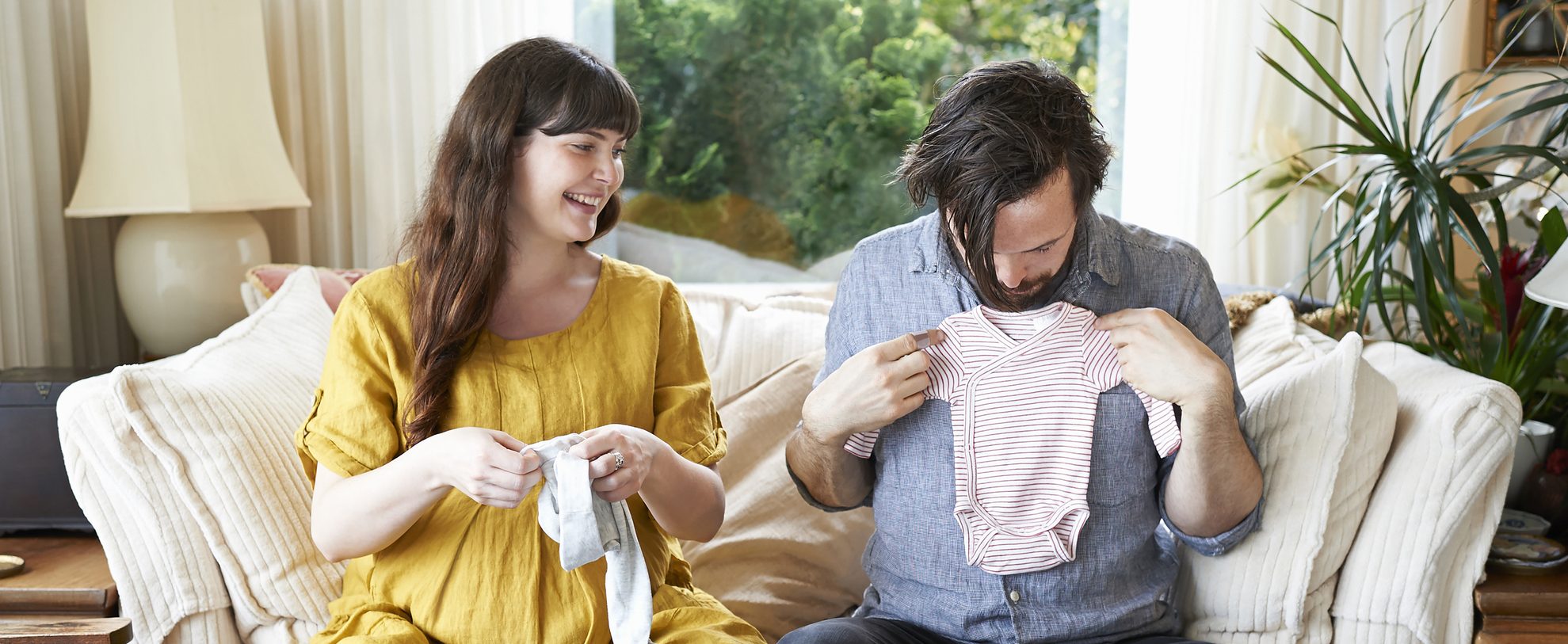 A man and a woman sit on a couch together, each holding some baby clothes. The man holds up a onesie to his chest.