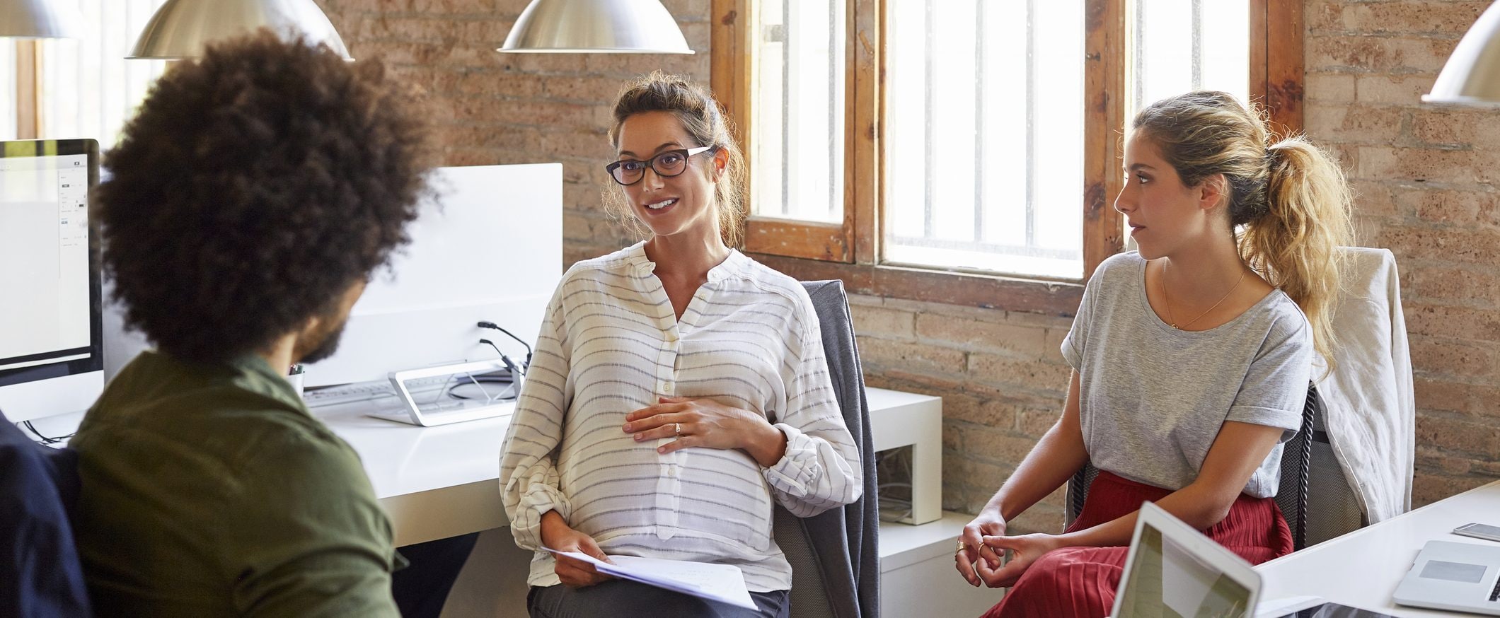 A young pregnant woman sits between two colleagues in an office setting.