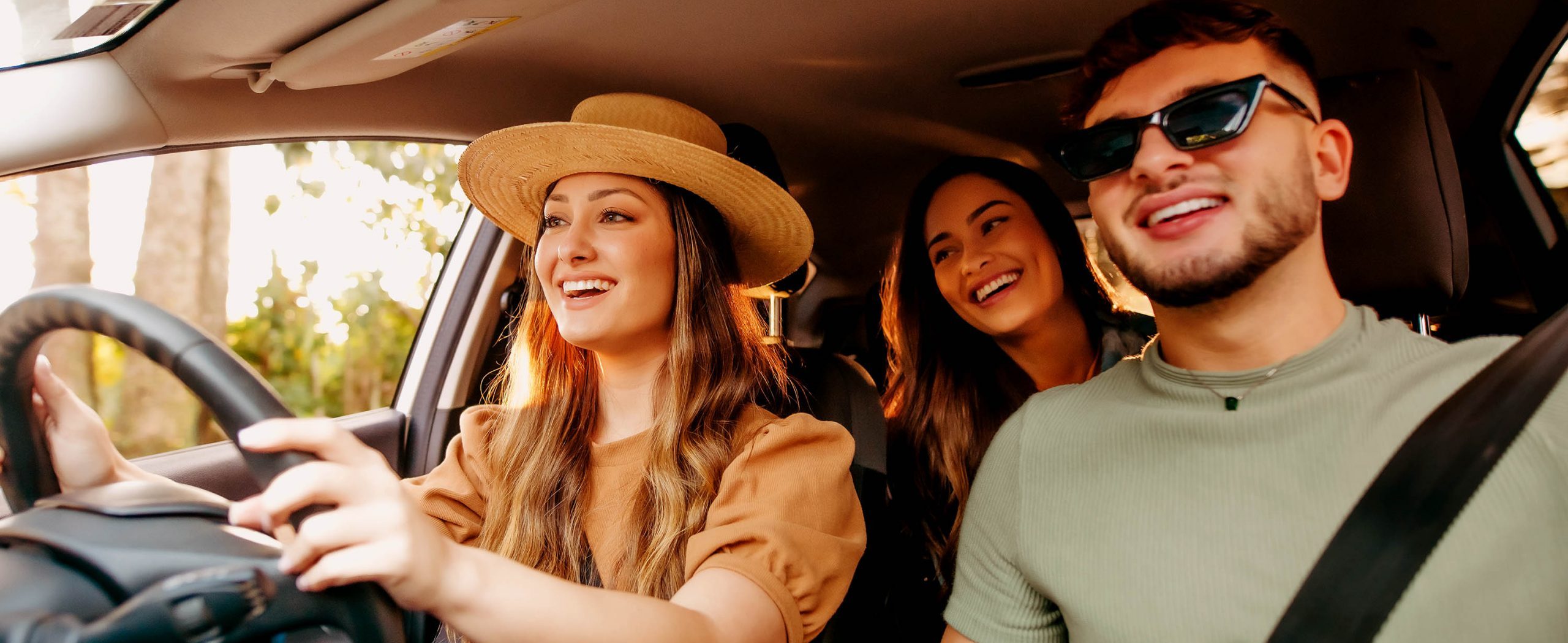 Two women and a man riding in a car, smiling, with trees in the background.