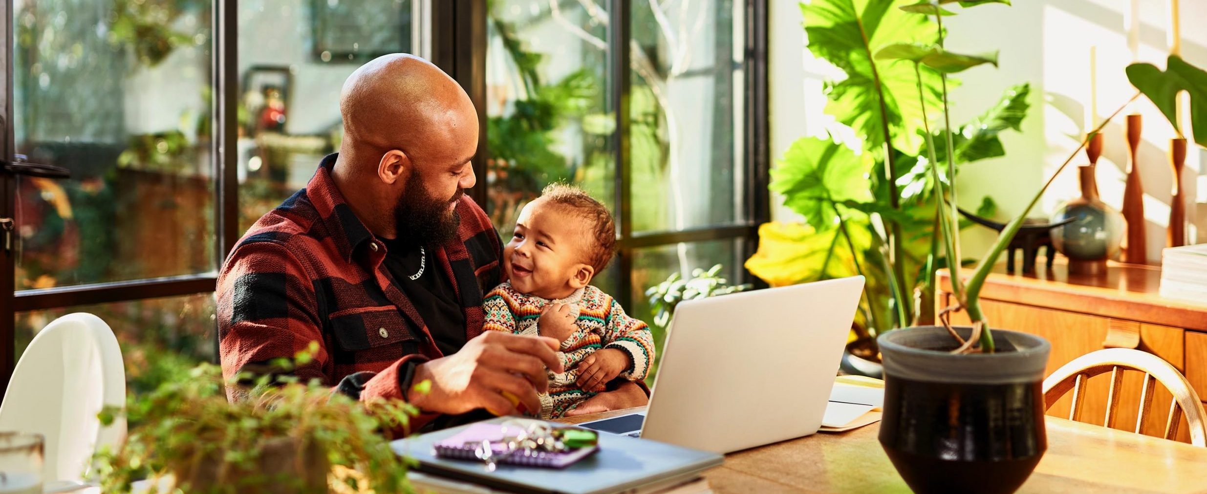 A man sits at a desk, smiling and holding a baby, next to plants and a laptop computer.