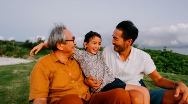 A grandpa, his son, and grandchild sit outside in the grass laughing.