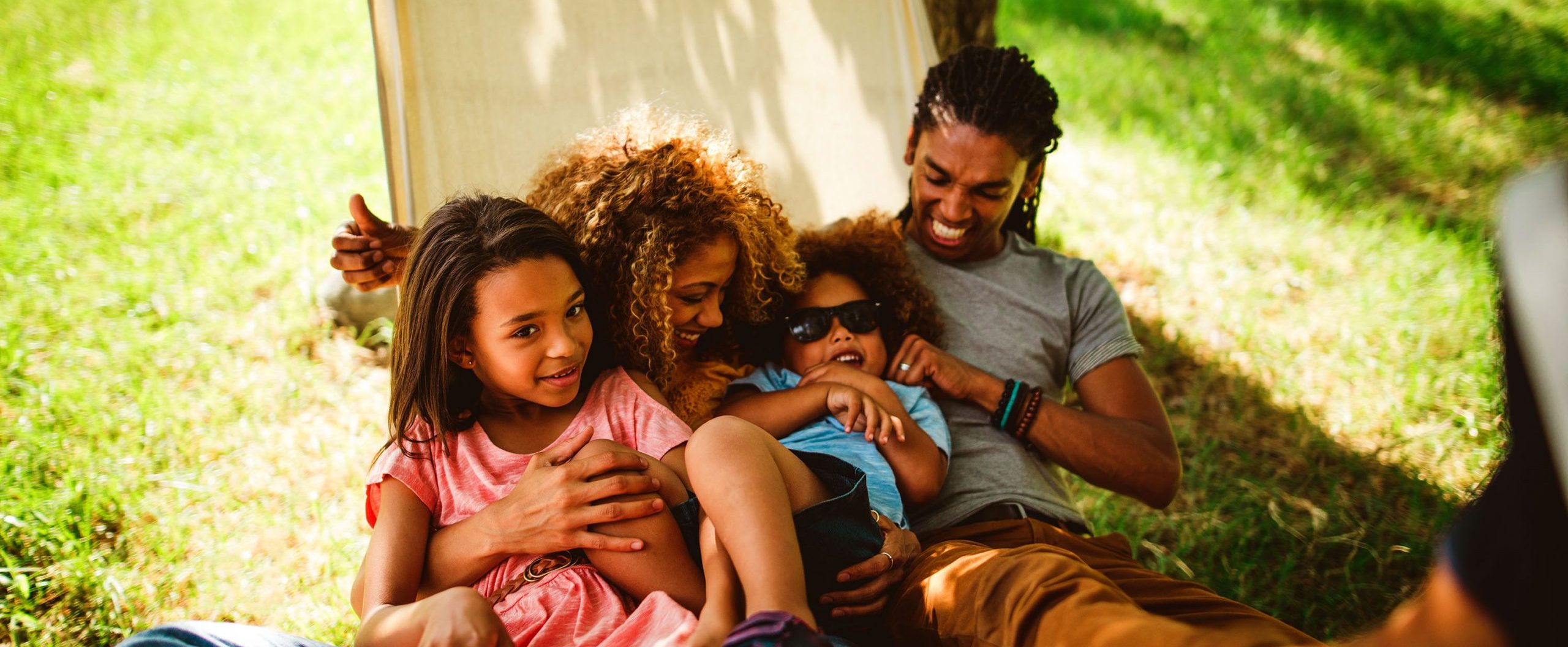 A family of four smiles while sitting in a hammock, with grass in the background.