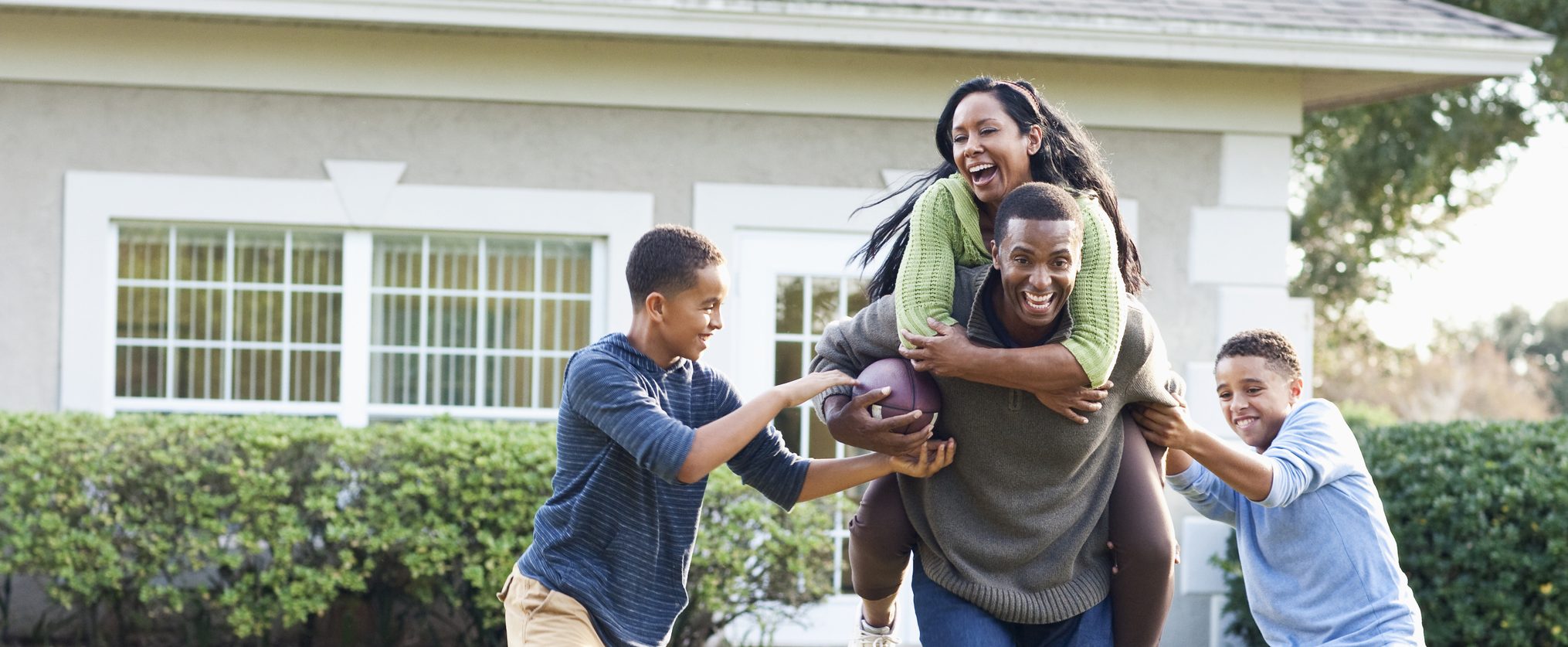 A family plays football outside. The dad gives the mom a piggyback ride and two sons are trying to get the ball and tackle them.