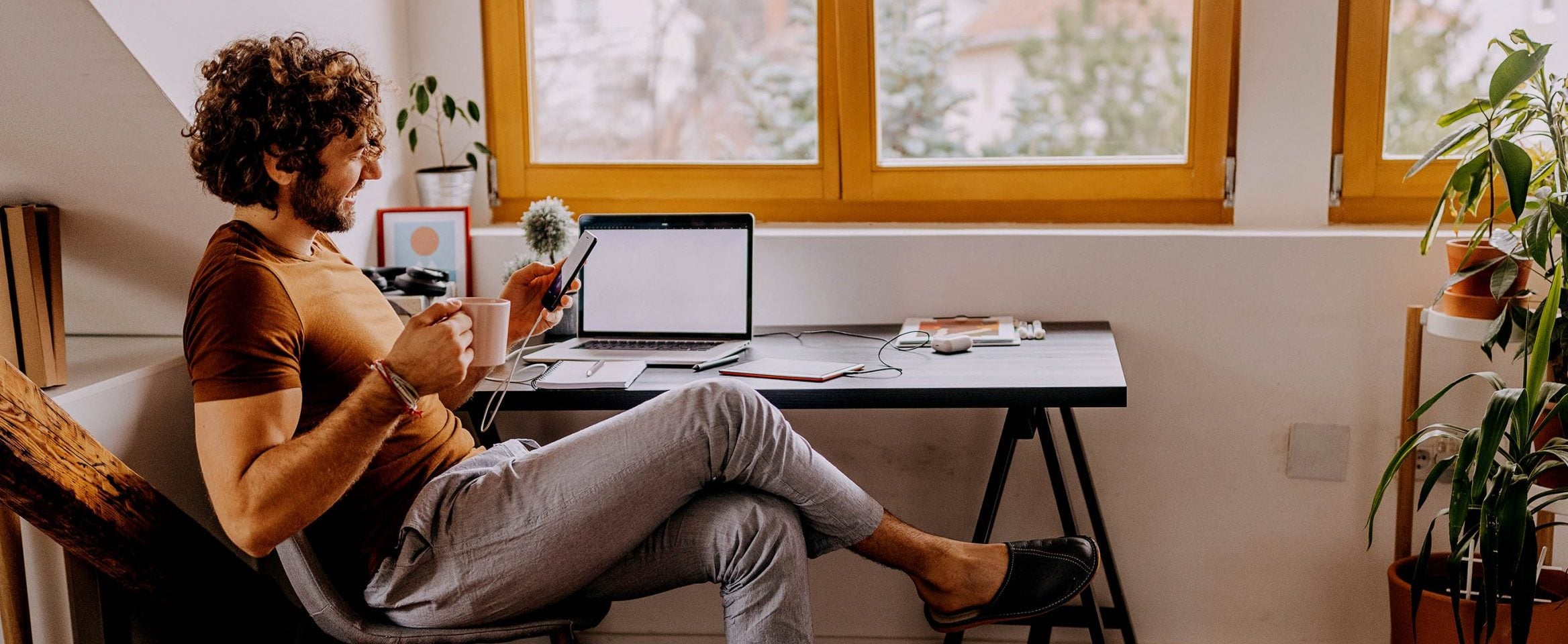 A young man smiles while holding a mug and looking at a mobile phone, next to a desk with laptop computer.