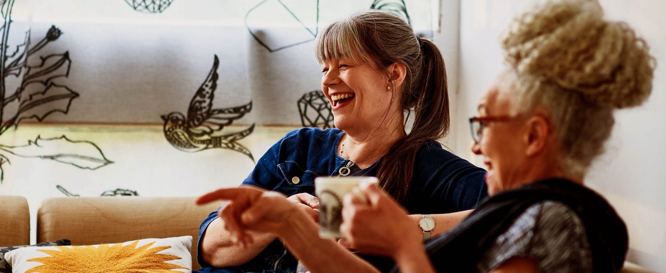 Two women sit on a sofa with patterned pillows, holding mugs and laughing.