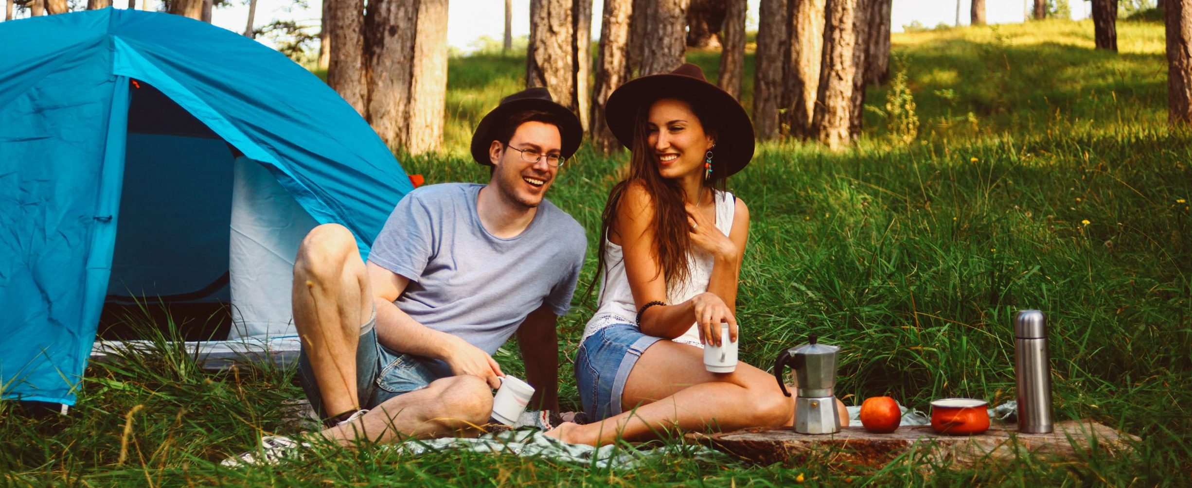 A man and woman hold mugs of coffee and smile while sitting on grass in front of a pitched tent.
