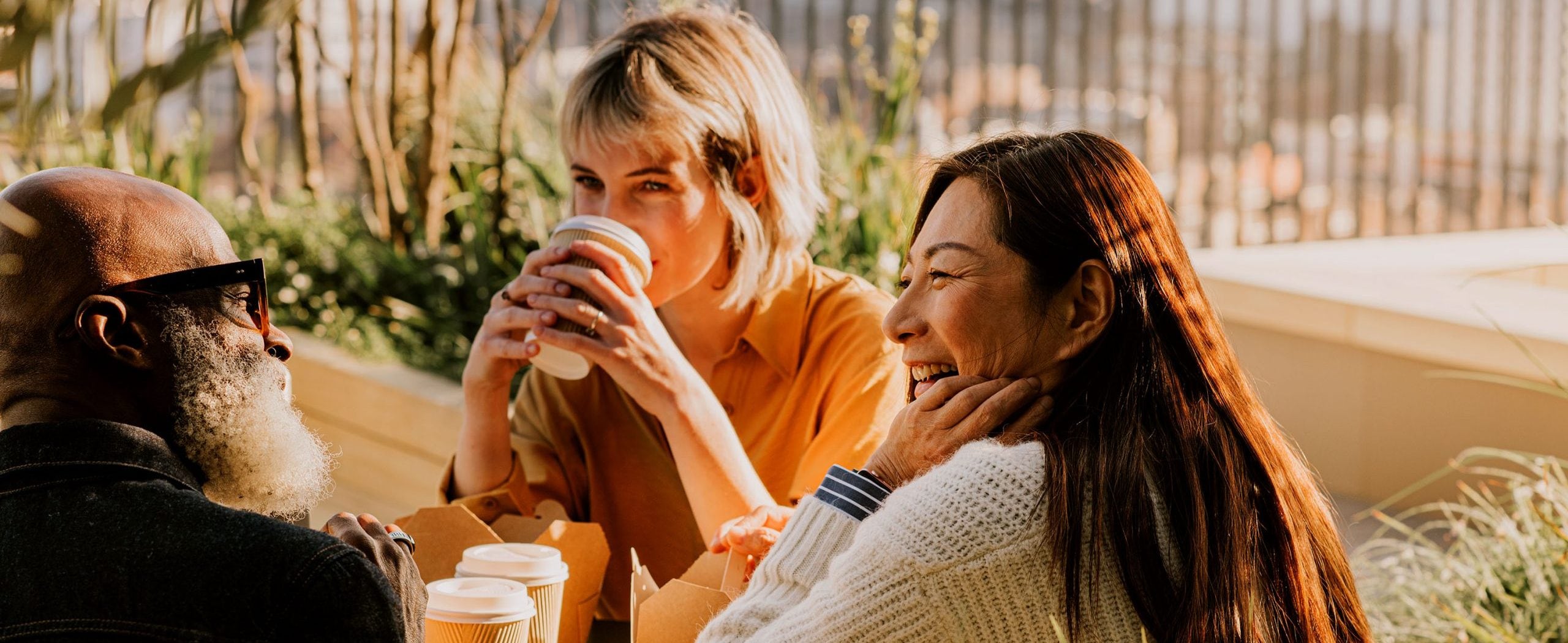 A man and two women smile and drink coffee at an outdoor table, with plants in the background.