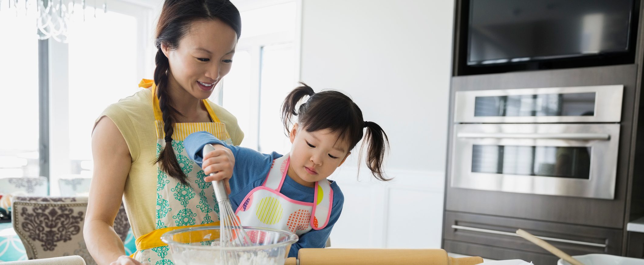 Caretaker oversees toddler mixing together flour with other ingredients in a kitchen.