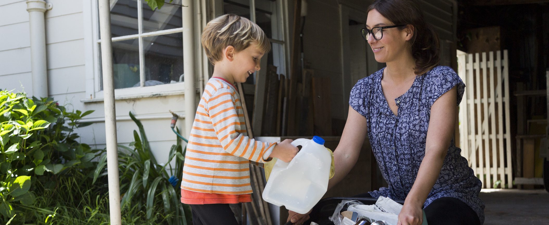A little kid is holding an empty plastic milk jug while a smiling woman holds a box of recycled products.