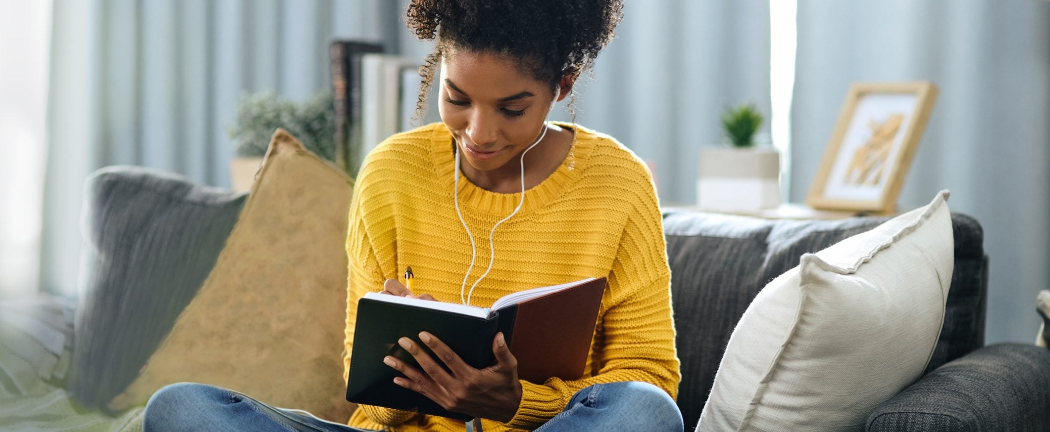 A woman in a yellow sweater sits cross legged on a couch writing in a journal and listening to something through ear buds.