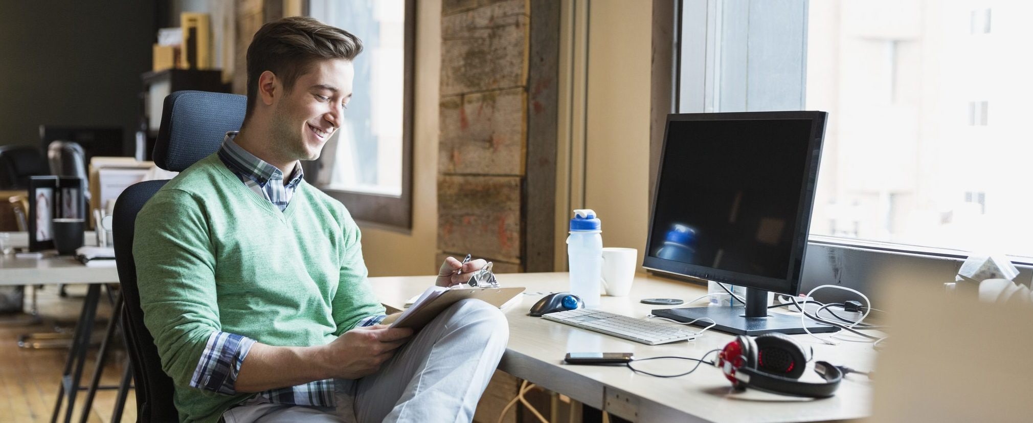 A man sits at a computer in an office smiling as he fills out some paperwork on a clipboard.