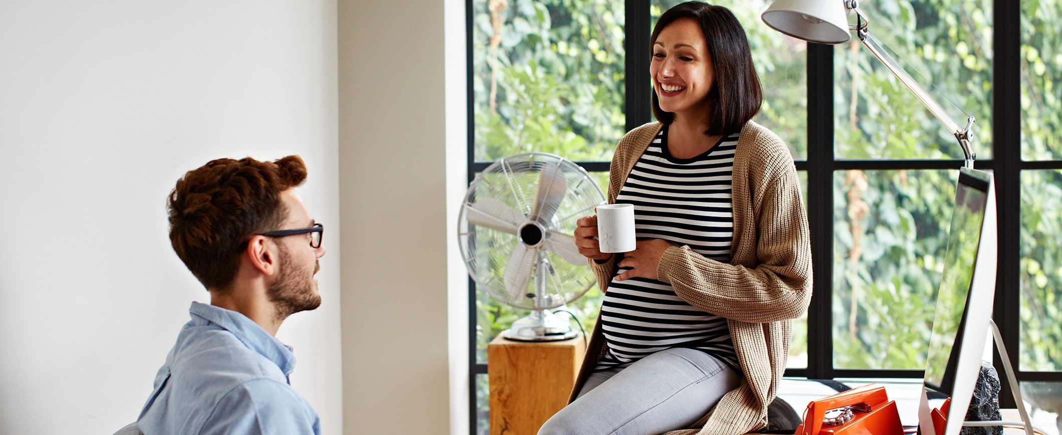 A pregnant woman sits on a desk with a mug in her hand, talking to a man sitting in a desk chair.