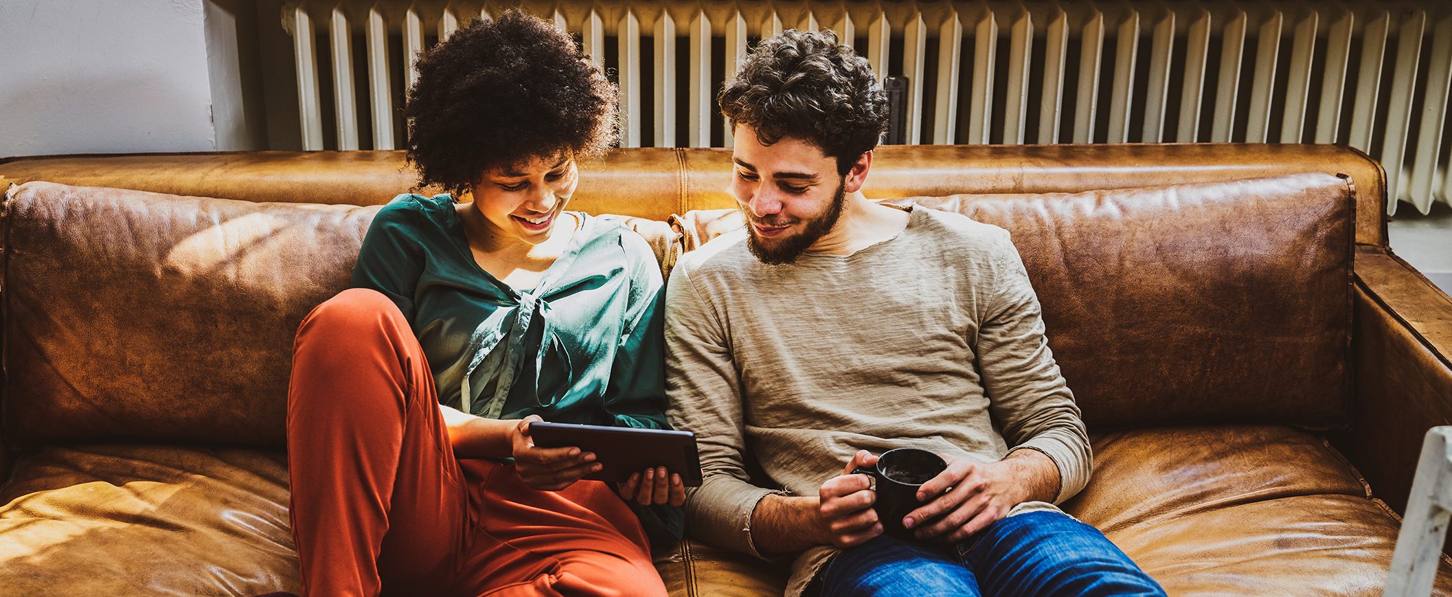 A man and woman sit together on a brown leather couch looking at a tablet together.