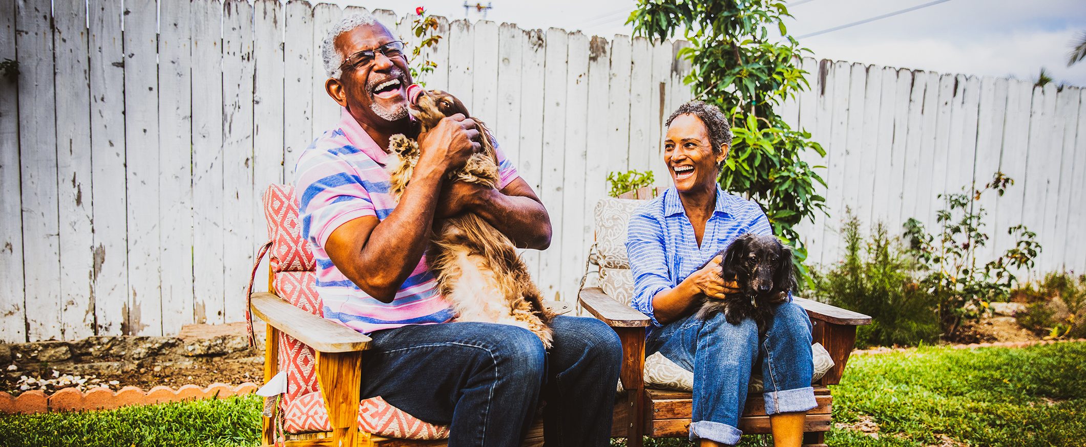 A man and a woman sit outside on chairs, each holding a puppy.