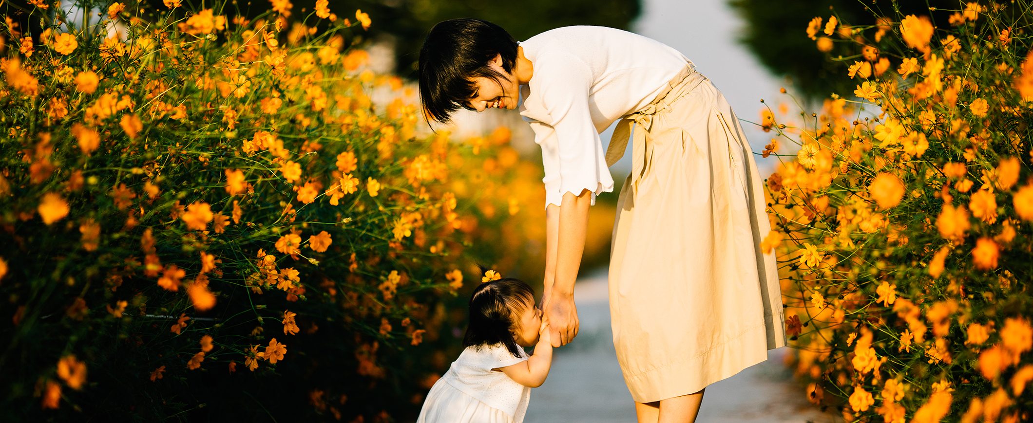 A woman bends down and holds the hands of a toddler in the middle of a path that is lined on either side with flowers.