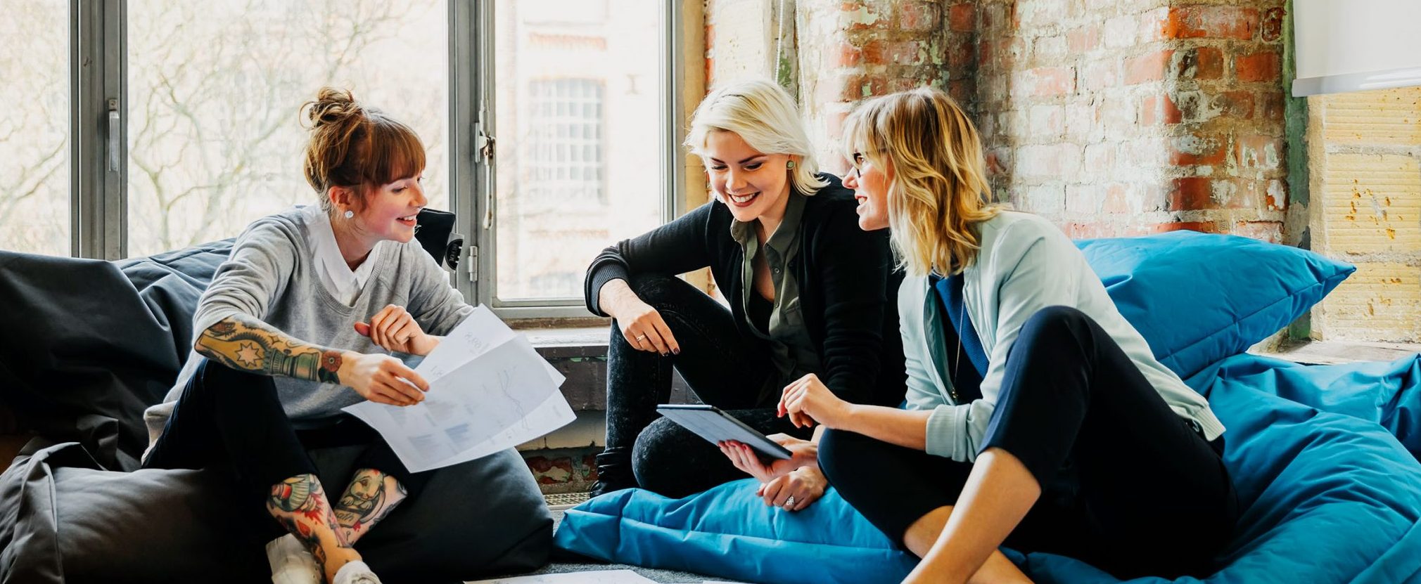 Three women sitting on sofas and talking, one holding a tablet, with windows and a brick wall in the background.