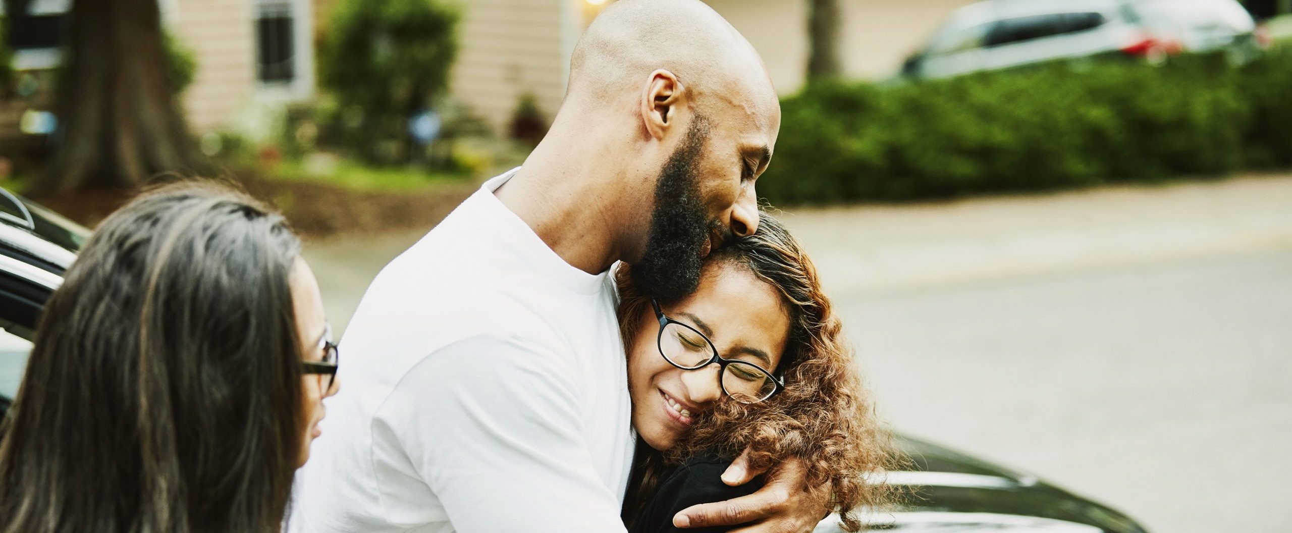 A man and woman smile and embrace as another woman looks on, with a car in the background.