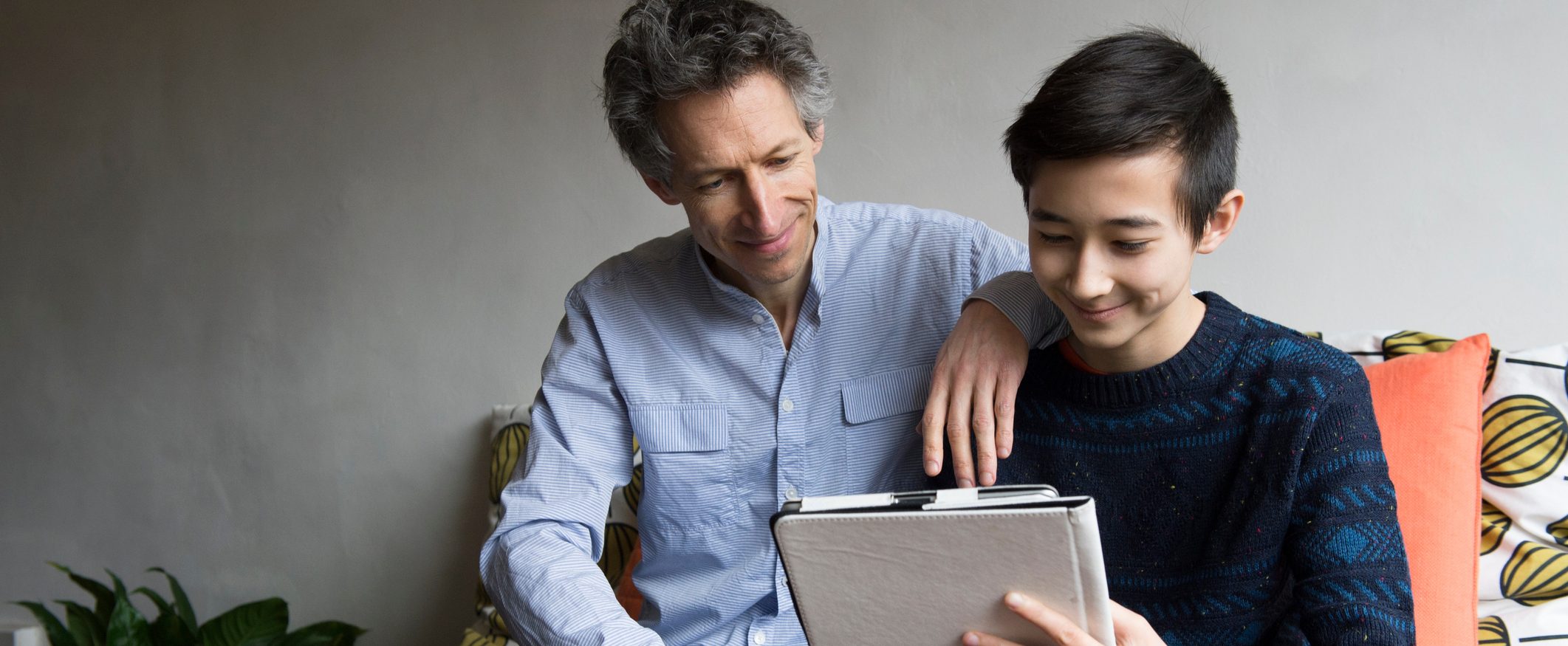A dad and son sit together while the son works on a tablet. The dad rests his arm on his son’s shoulder.