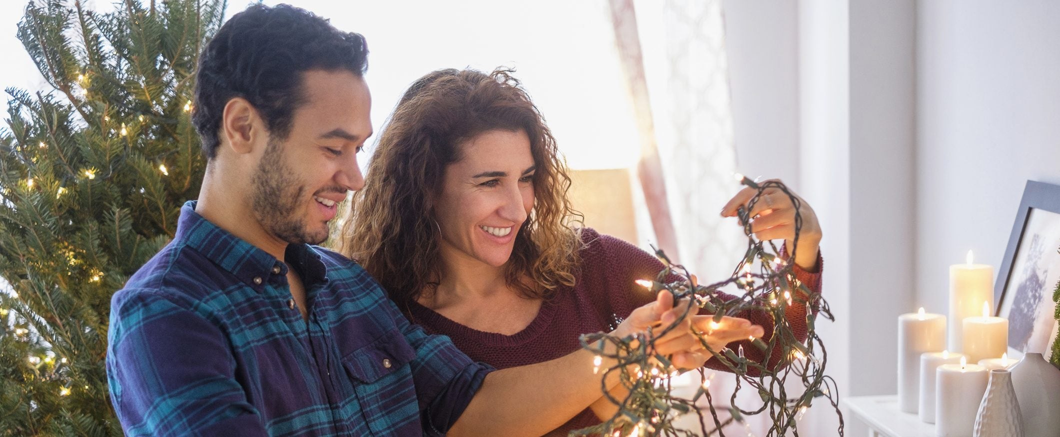 A man and woman stand in front of a Christmas tree holding a tangle of Christmas lights.