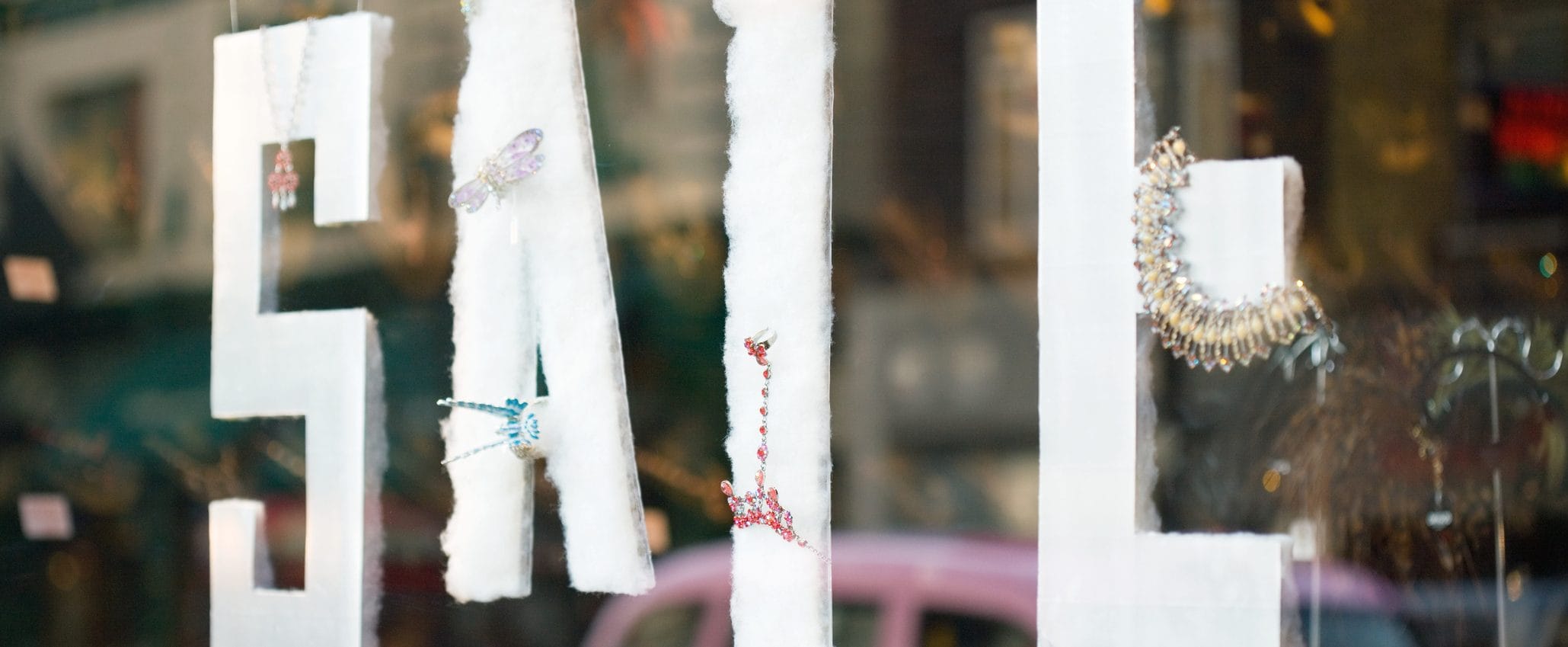 A shop window with the letters “SALE” hanging inside. There are different pieces of jewelry adorning the letters.