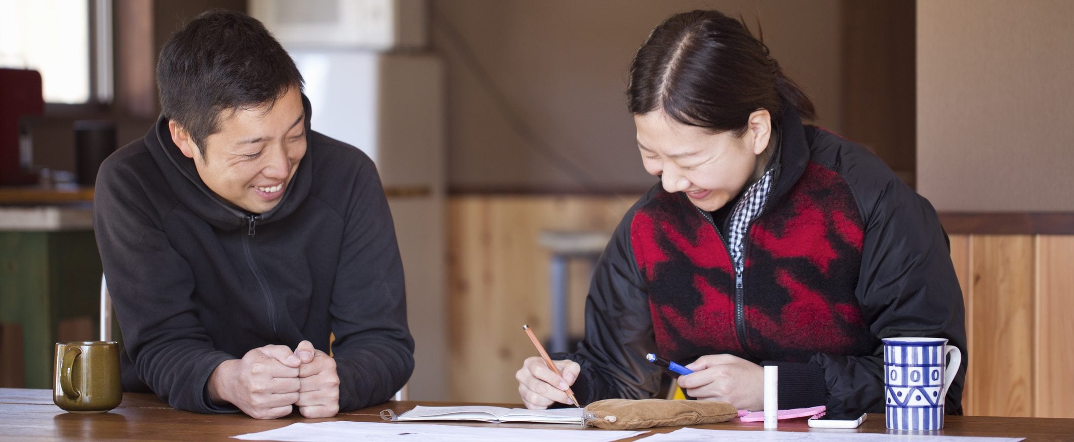 Two people sit at a table looking at some papers. There are also mugs on the table.