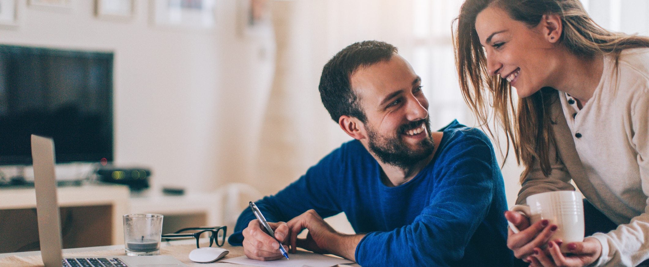 A man sitting at a desk writing something on a piece of paper looks up at a woman holding a coffee cup and smiles.