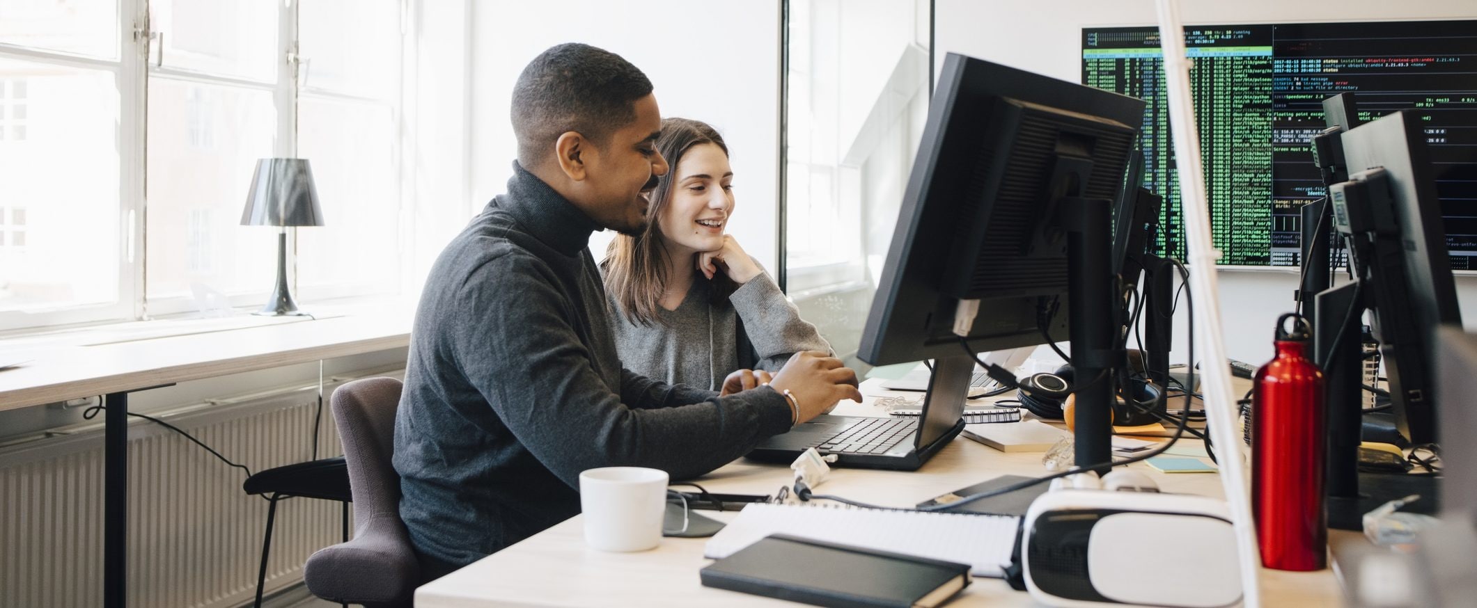 Two people sit together in an office environment and look at a computer monitor together.