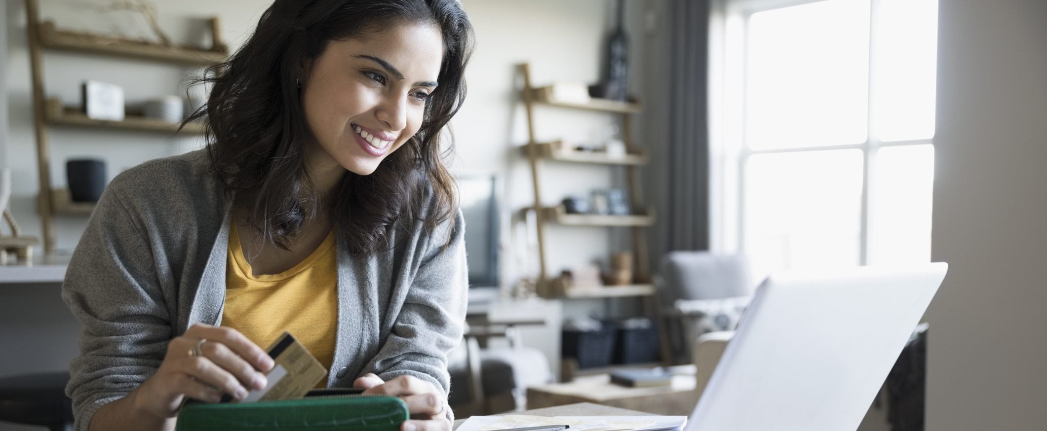 A woman smiles into the screen of a laptop while taking a credit card out of her wallet.