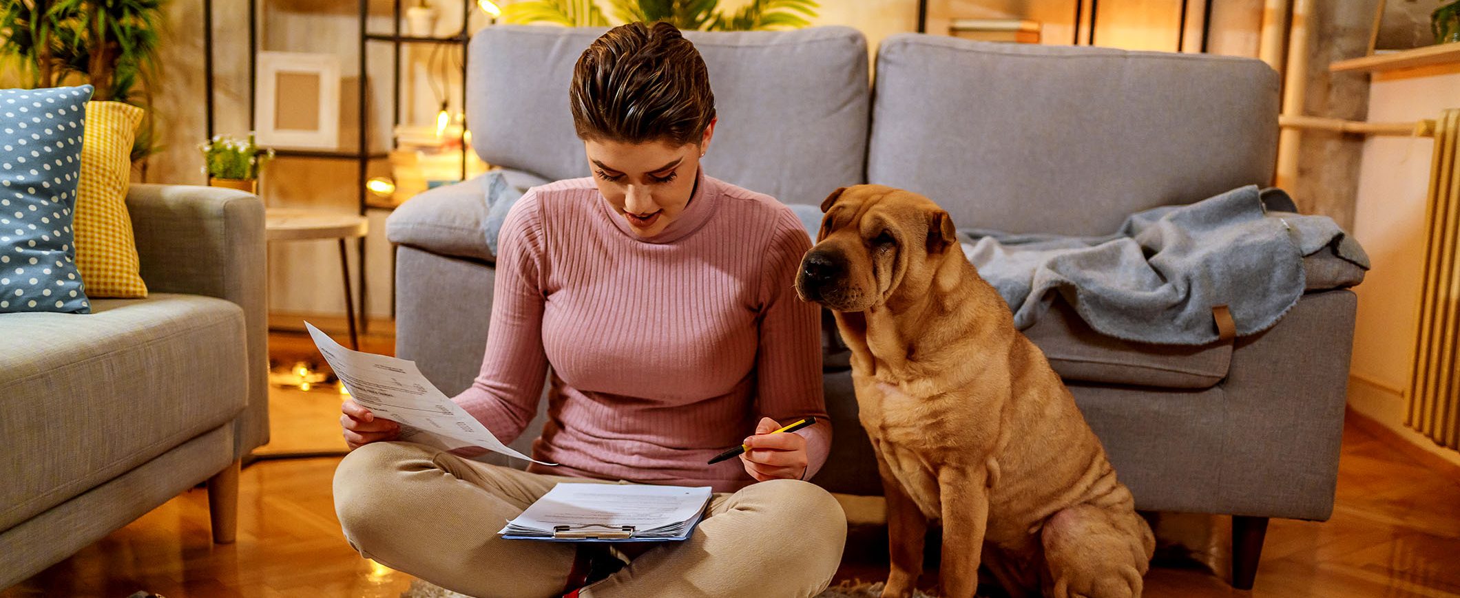 A woman sits with her dog on the floor of her living room, reviewing documents.