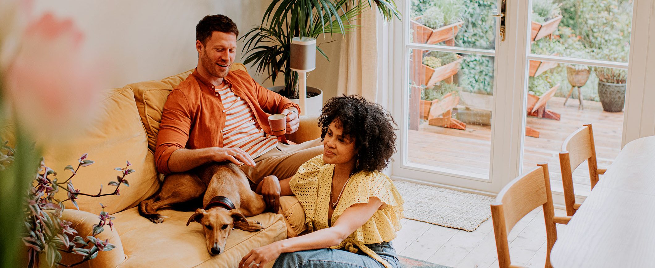 A man and woman relax with their dog on a sofa, with glass doors and plants in the background.