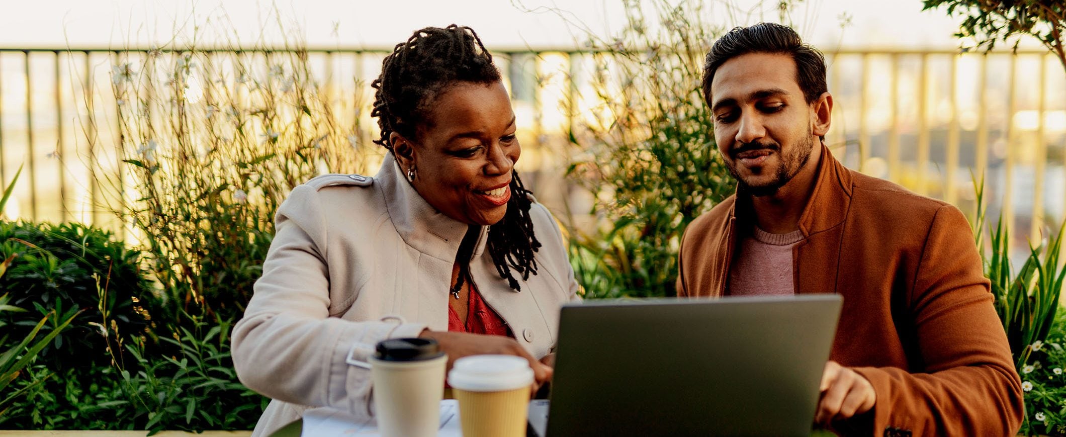 A man and woman sit at a table outdoors with a laptop and cups of coffee.