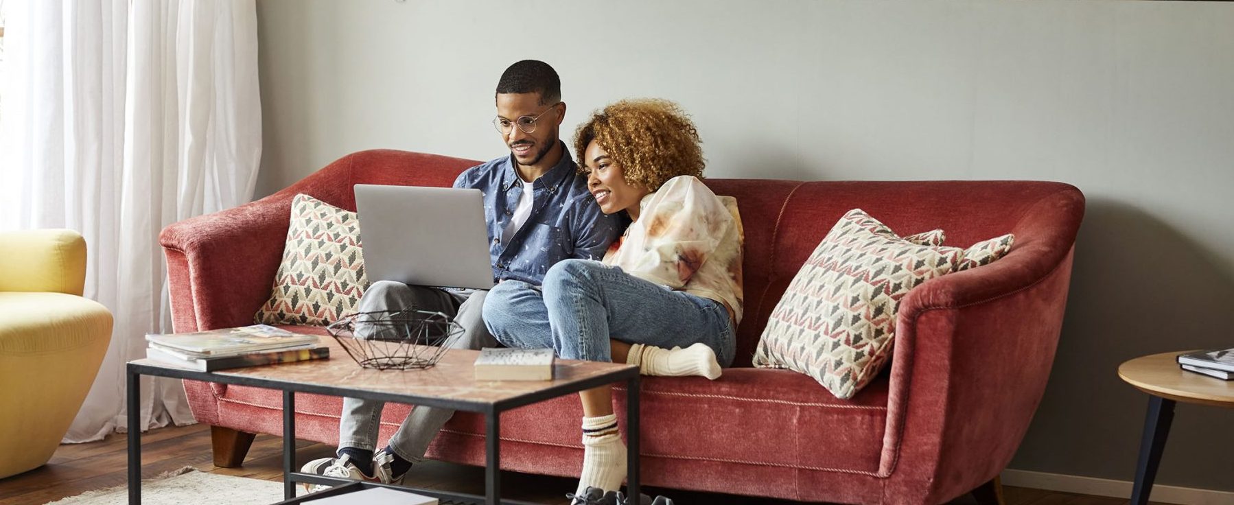 A couple sit together on a red velvet couch and smile while watching a laptop.