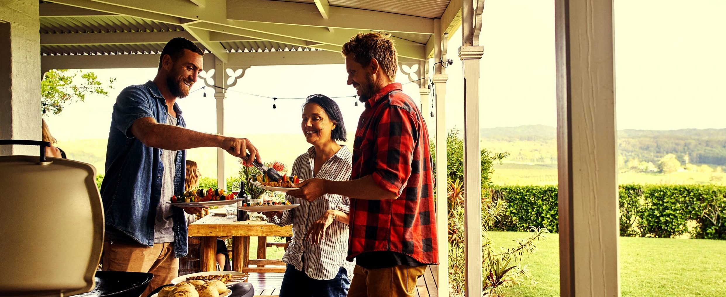 Three friends enjoy plates of grilled food on a sunny porch, with plants in the background